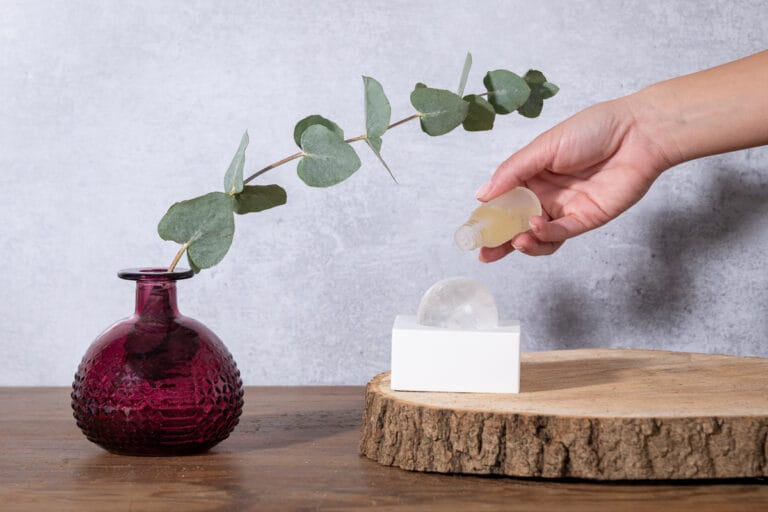 Hand model holding a diffusing white block with one hand and drawing a plant on it with the other, with a sphere of precious rock usually embedded in the block standing next to it. She is now dropping essential oils onto the rock, creating a serene and artistic scene that showcases the elegant design and natural elements of the diffuser.
