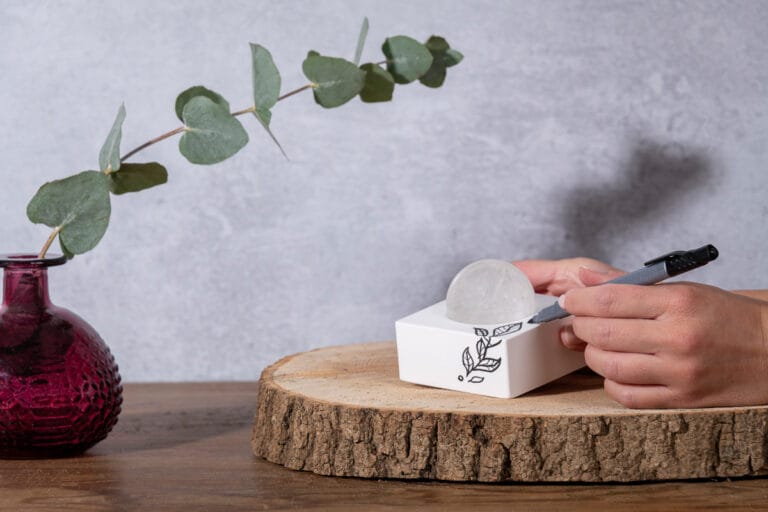 Hand model holding a diffusing white block with one hand and drawing a plant on it with the other, with a sphere of precious rock that is embedded in the block, creating a serene and artistic scene that showcases the elegant design and natural elements of the diffuser.