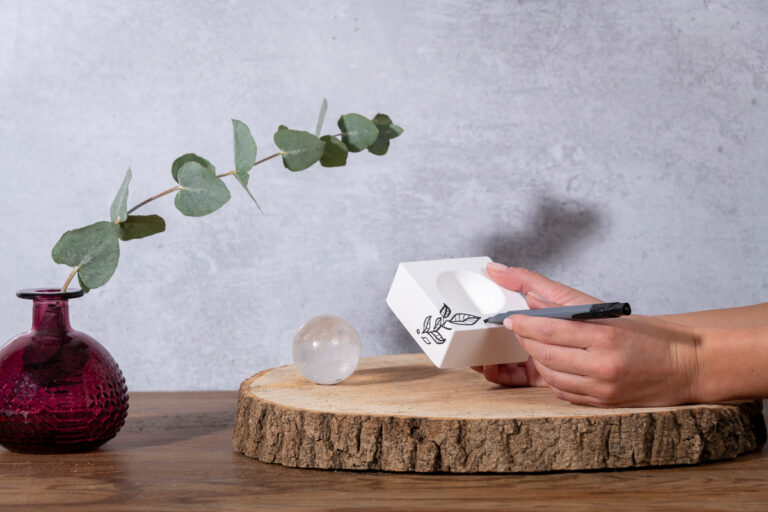 Hand model holding a diffusing white block with one hand and drawing a plant on it with the other, with a sphere of precious rock thats usually embedded in the block standing next to it, creating a serene and artistic scene that showcases the elegant design and natural elements of the diffuser.