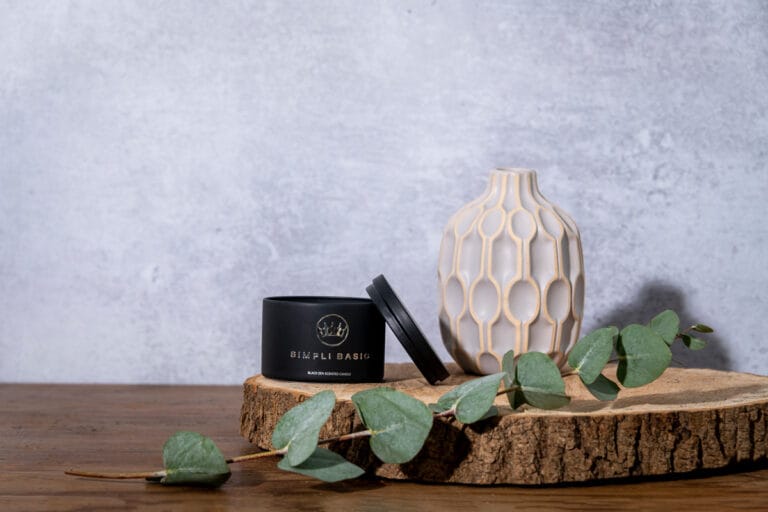 Candle with lid leaning on it, placed on a round of wood with a vase and foliage, creating a calm and stylish scene with natural elements.